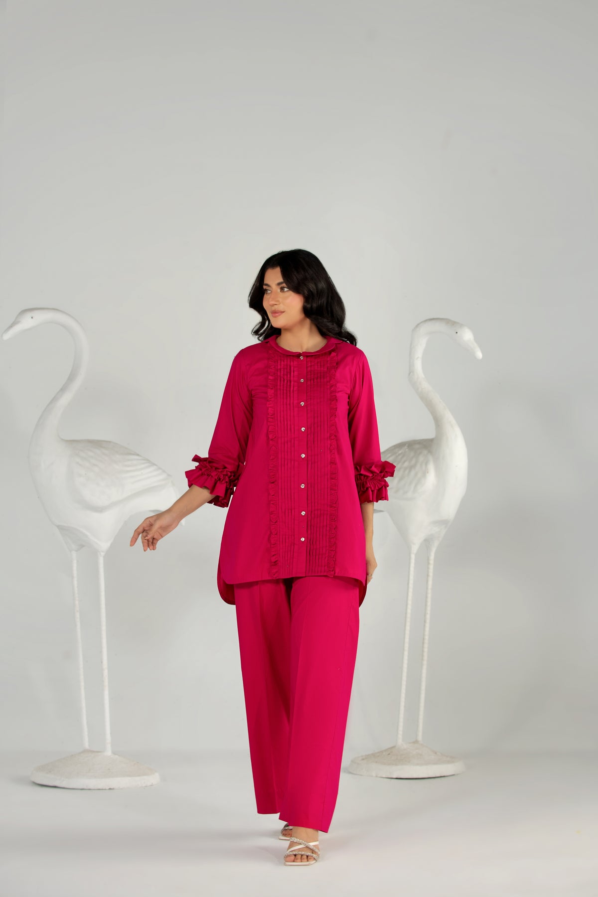 Woman wearing a bright pink outfit standing between two white chairs on a plain background