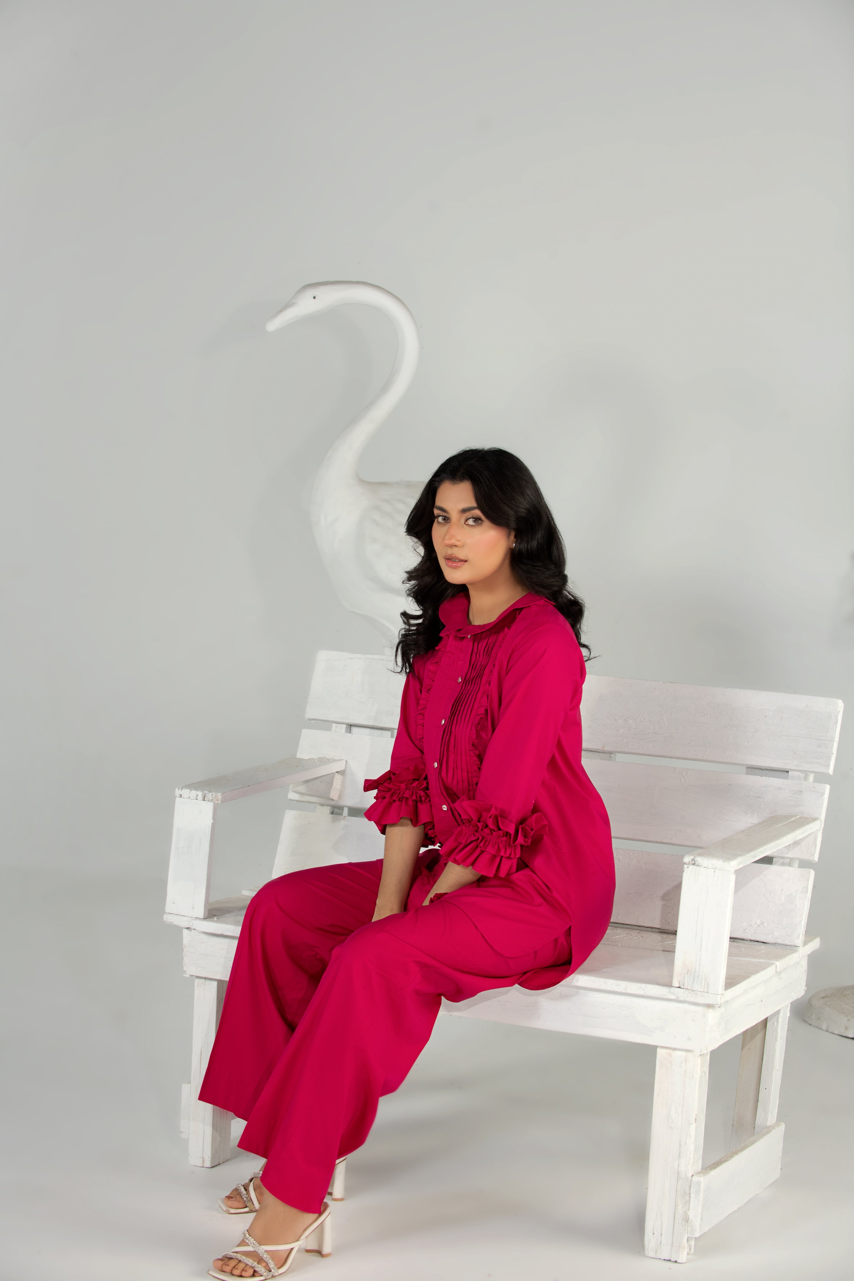 Woman in a bright pink outfit sitting on a white bench with a white swan sculpture in the background.