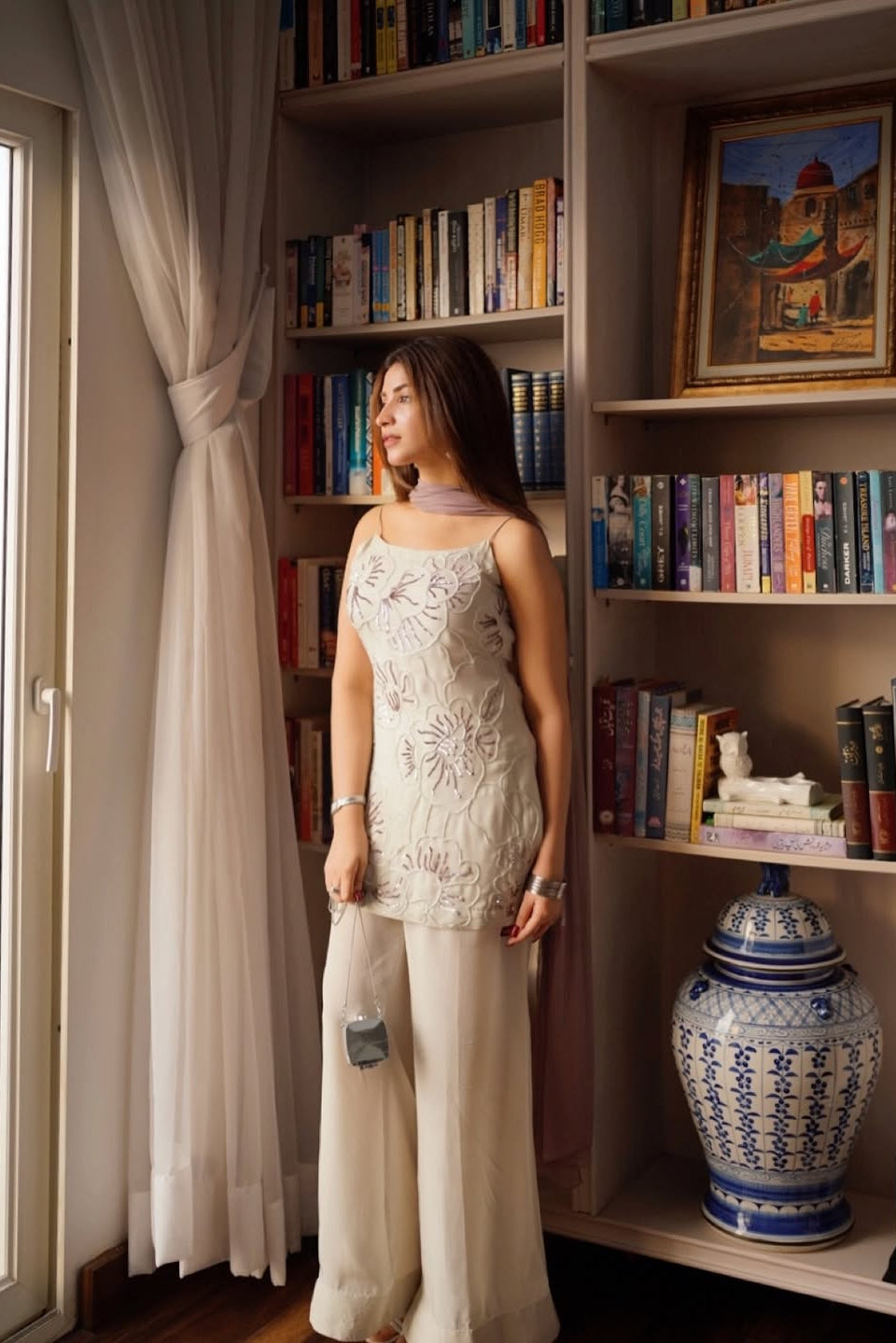 Woman in a white dress standing in a room with bookshelves and a large window.
