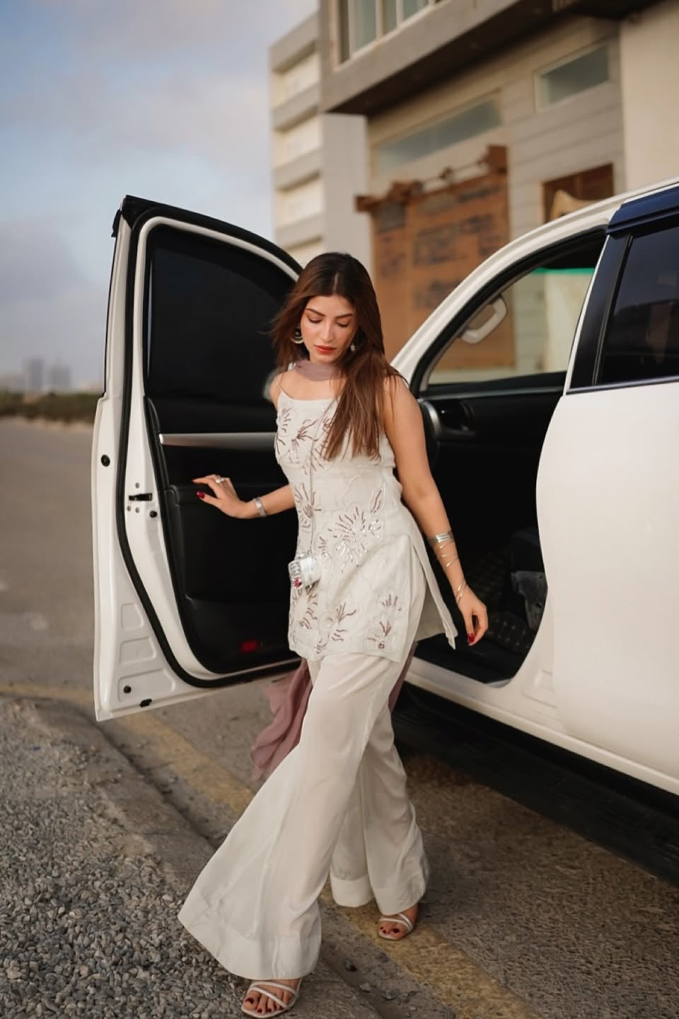 Woman in a white dress stepping out of a white car on a road.