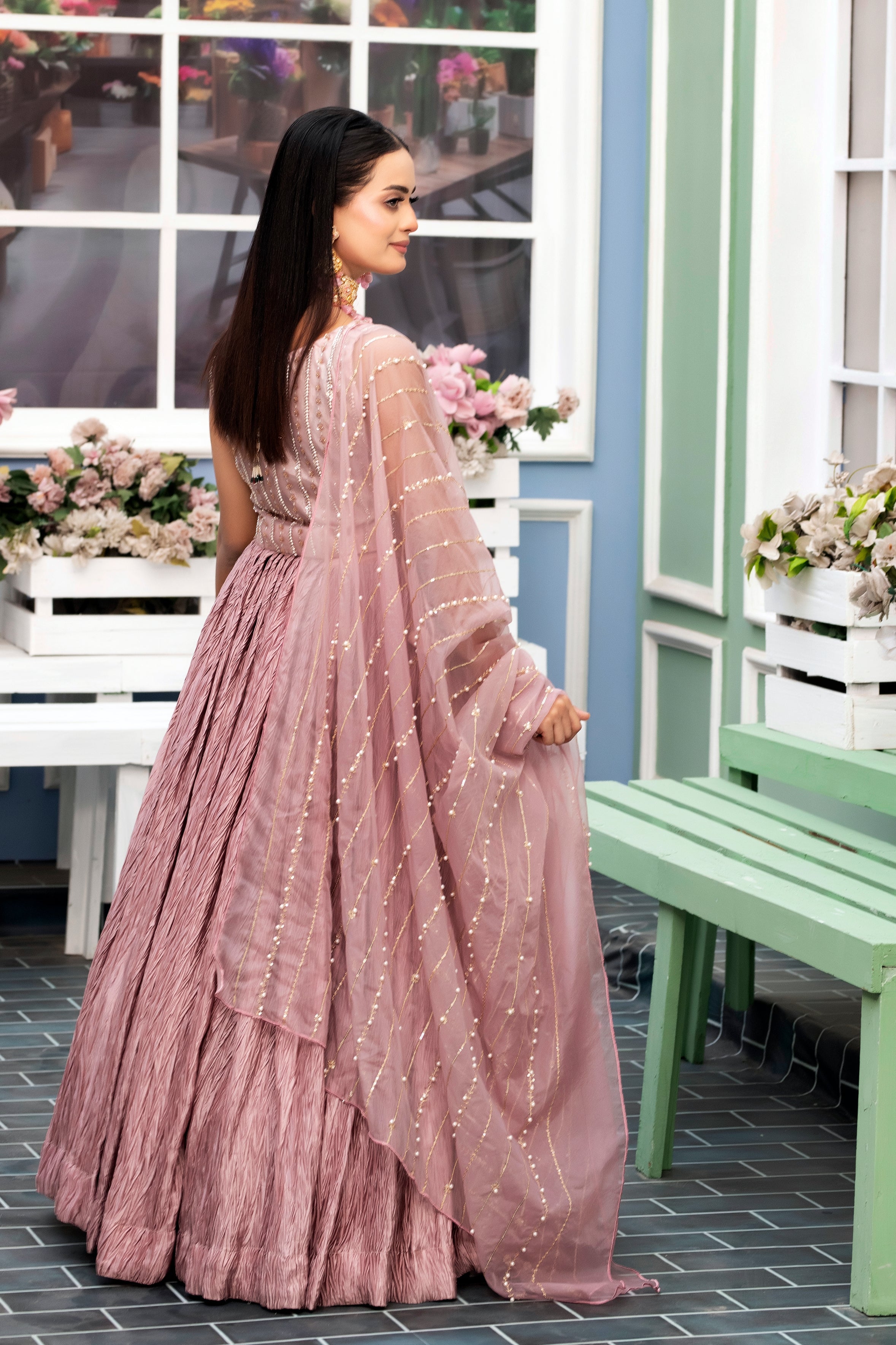 Woman in a pink traditional outfit standing in a decorative indoor setting with flowers and furniture.