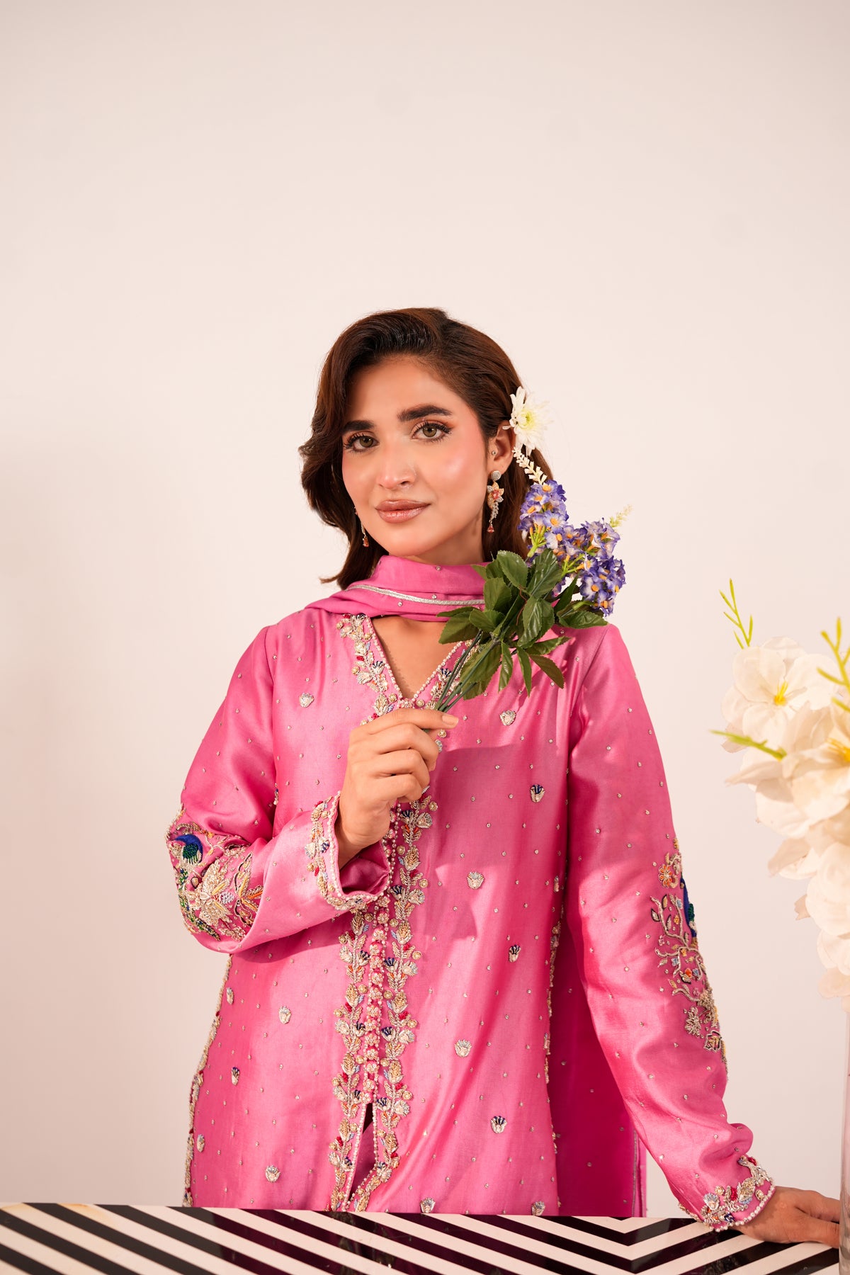 A woman wearing a bright pink embroidered traditional outfit with a matching scarf, holding a small bouquet of purple and white flowers against a plain light background.