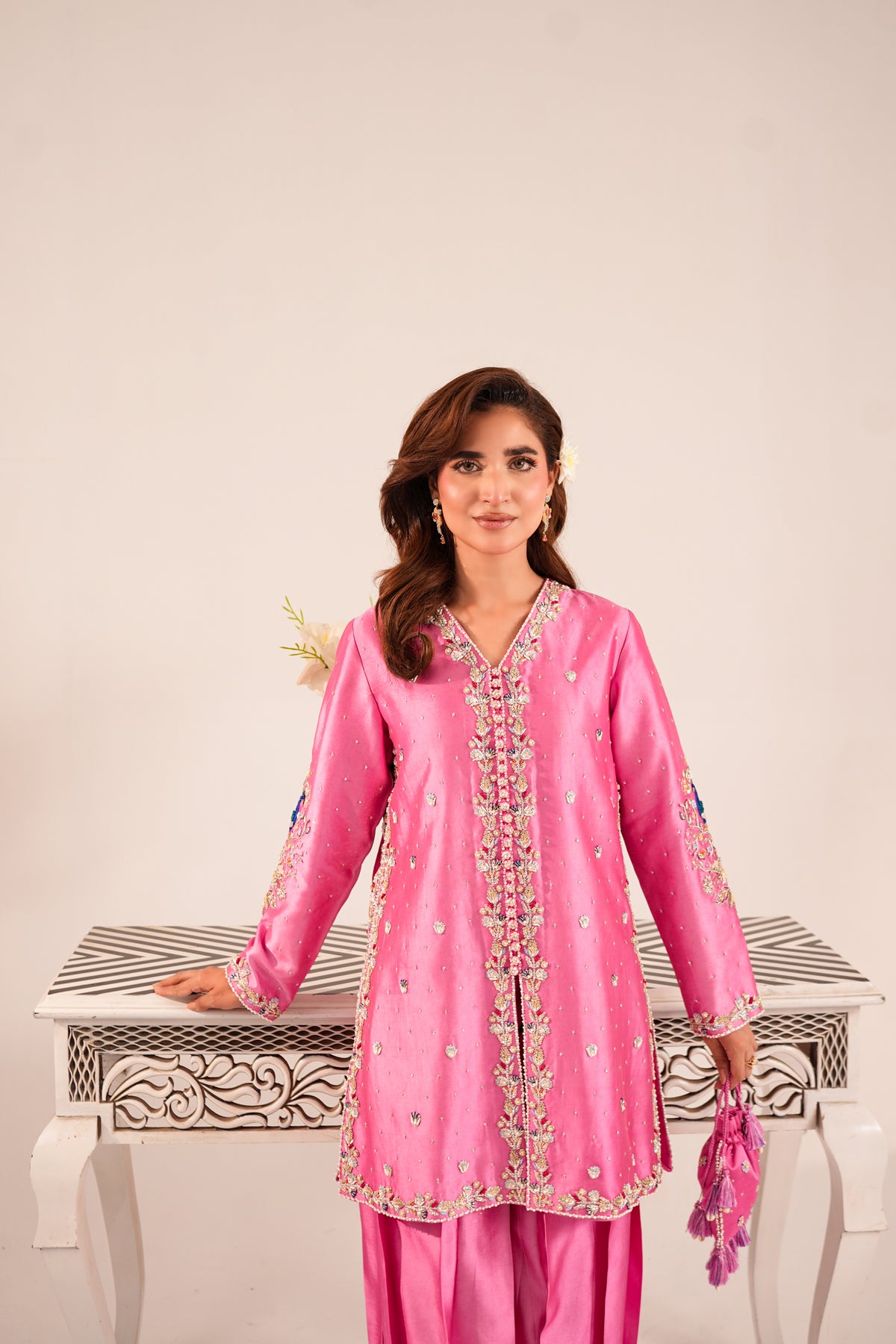 A woman modeling a bright pink traditional outfit with intricate gold and silver embroidery, standing behind a white decorative table against a plain beige background.