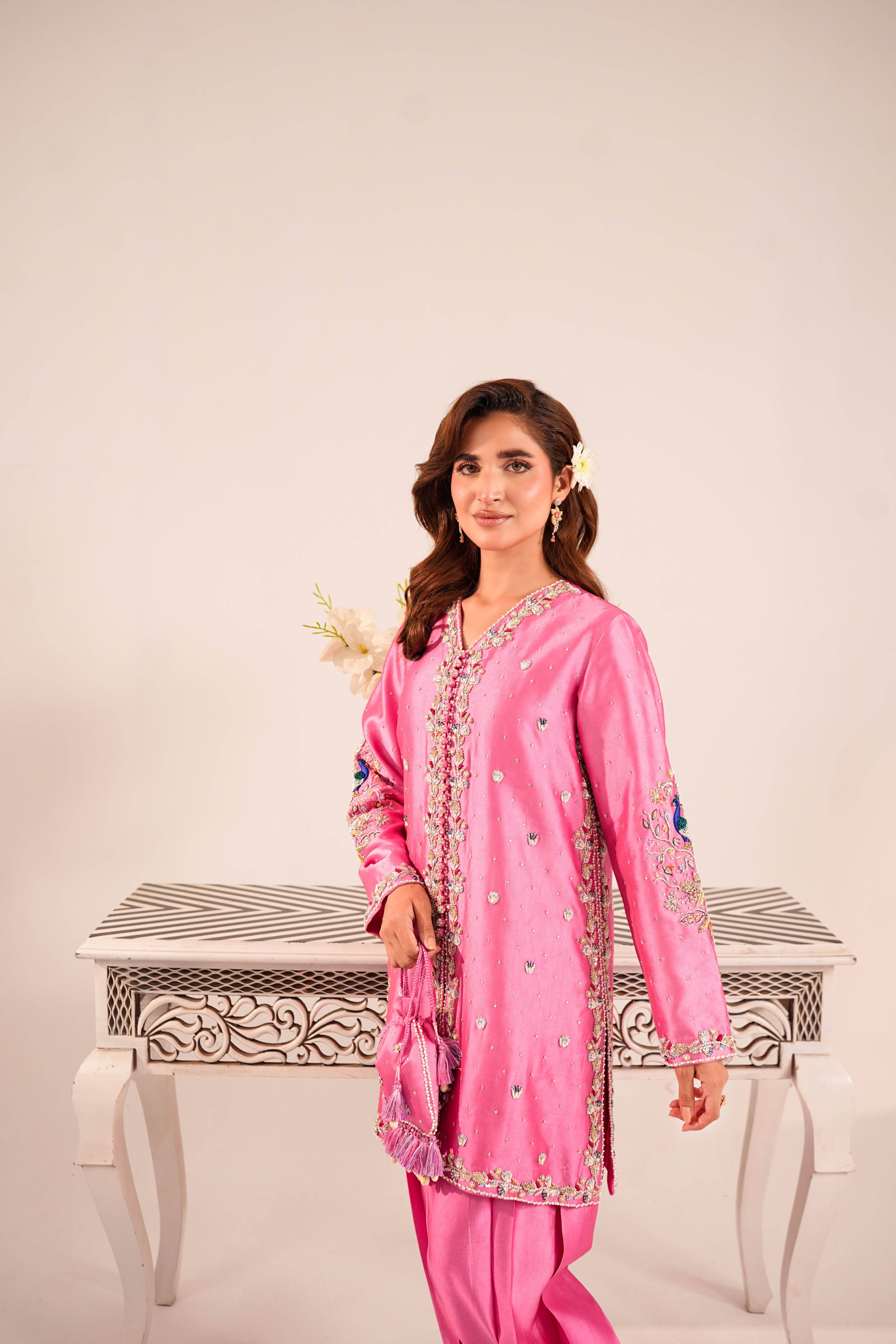 A woman modeling a pink traditional South Asian outfit with matching trousers, featuring intricate silver and gold embroidery along the neckline, sleeves, and hem, standing in front of a decorative white table against a light beige background.