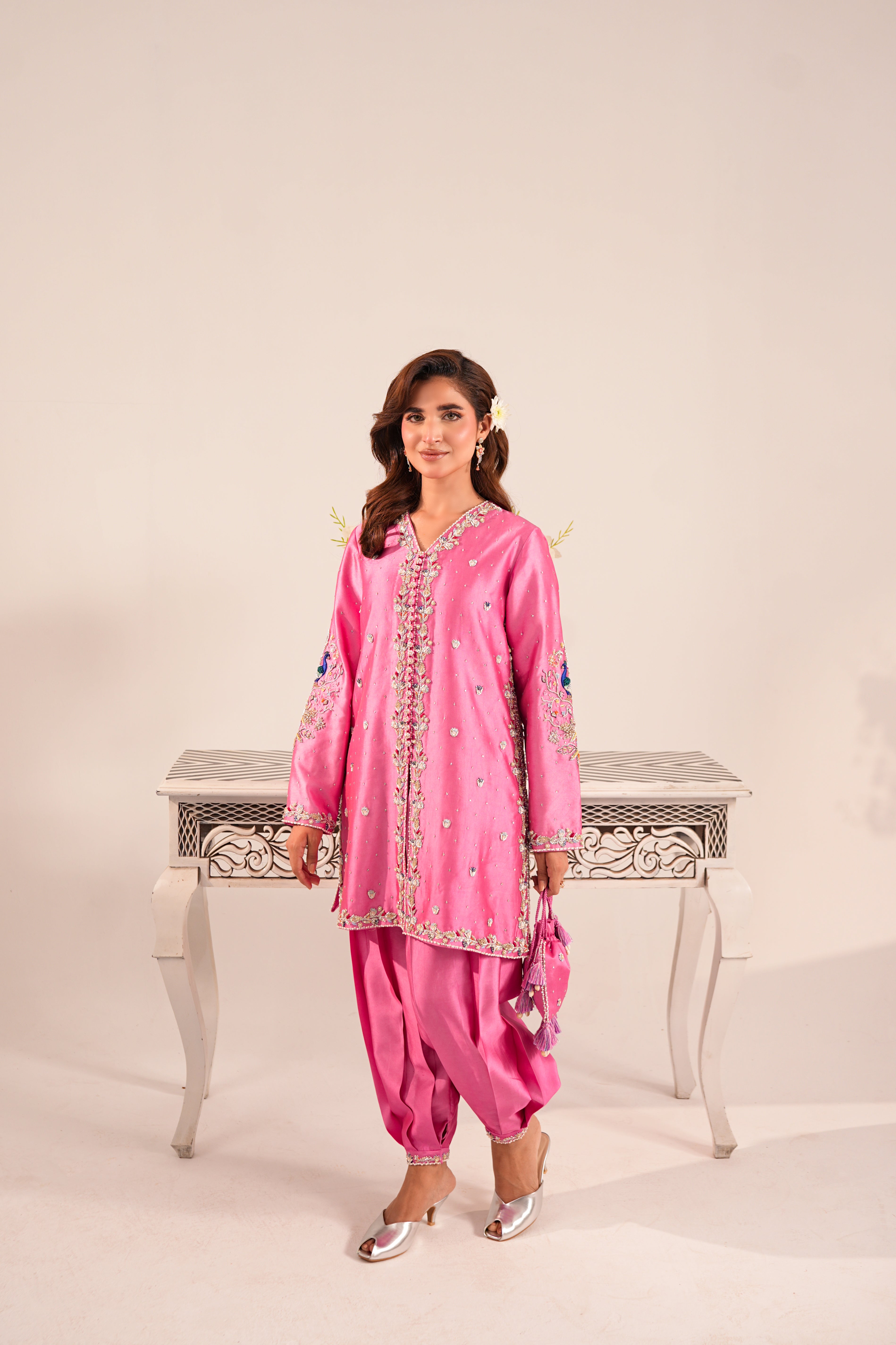 A woman modeling a pink traditional South Asian outfit with embroidered details, standing in front of a light beige studio backdrop and a white decorative table.