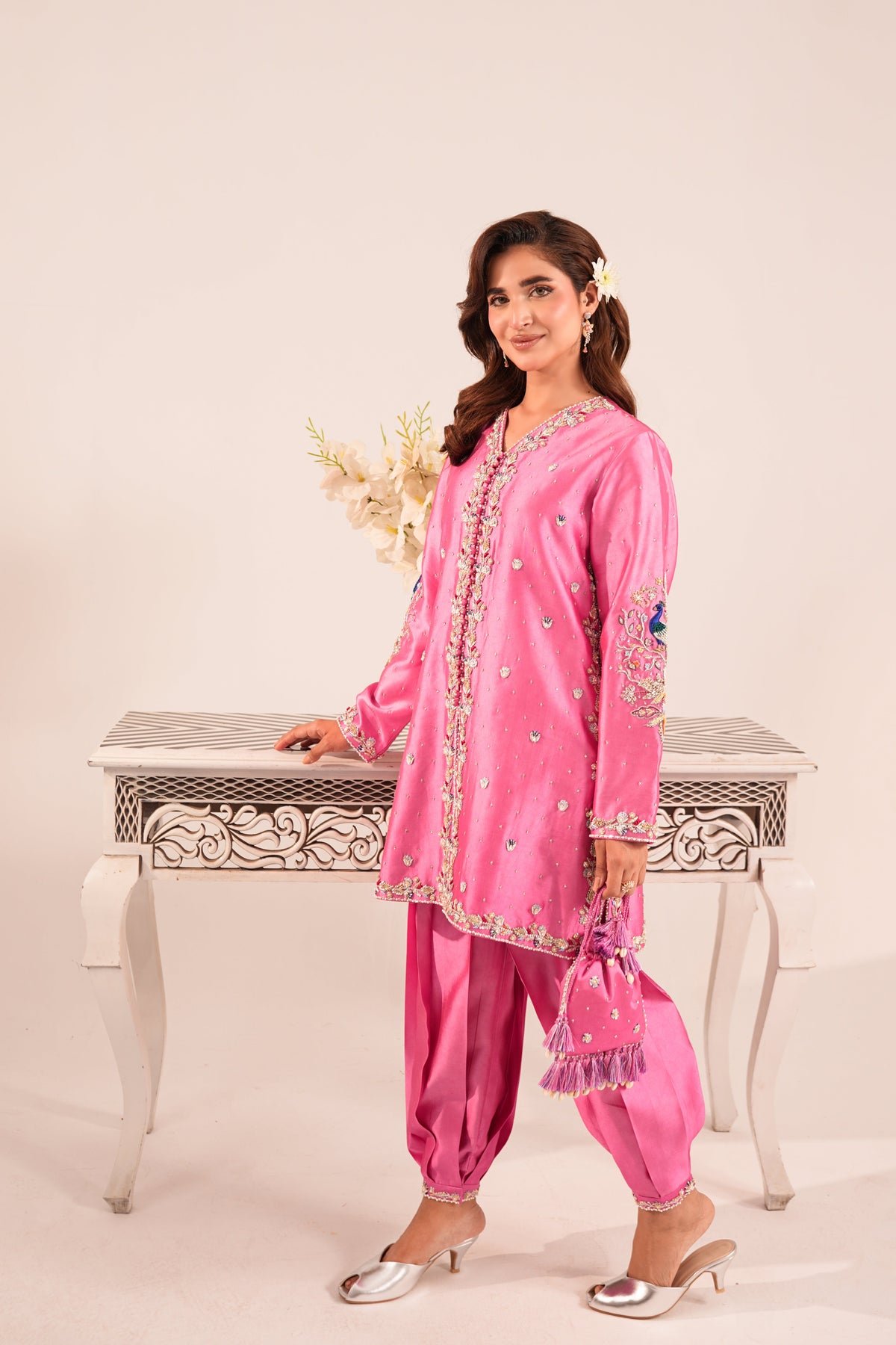A woman modeling a bright pink traditional South Asian outfit with matching pants, a long embroidered kurta, and silver heeled sandals, standing beside a decorative white table against a plain light beige studio background.