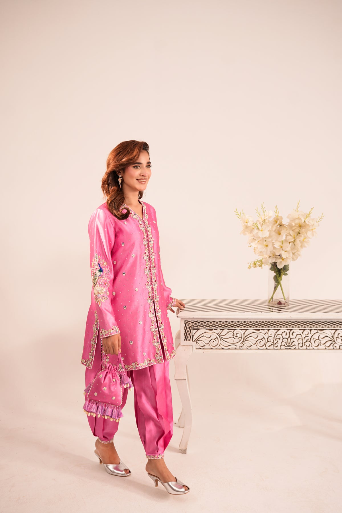A woman models a bright pink traditional South Asian outfit with matching pants, a long embroidered kurta, and metallic silver heels, standing next to a decorative table with flowers against a plain beige background.