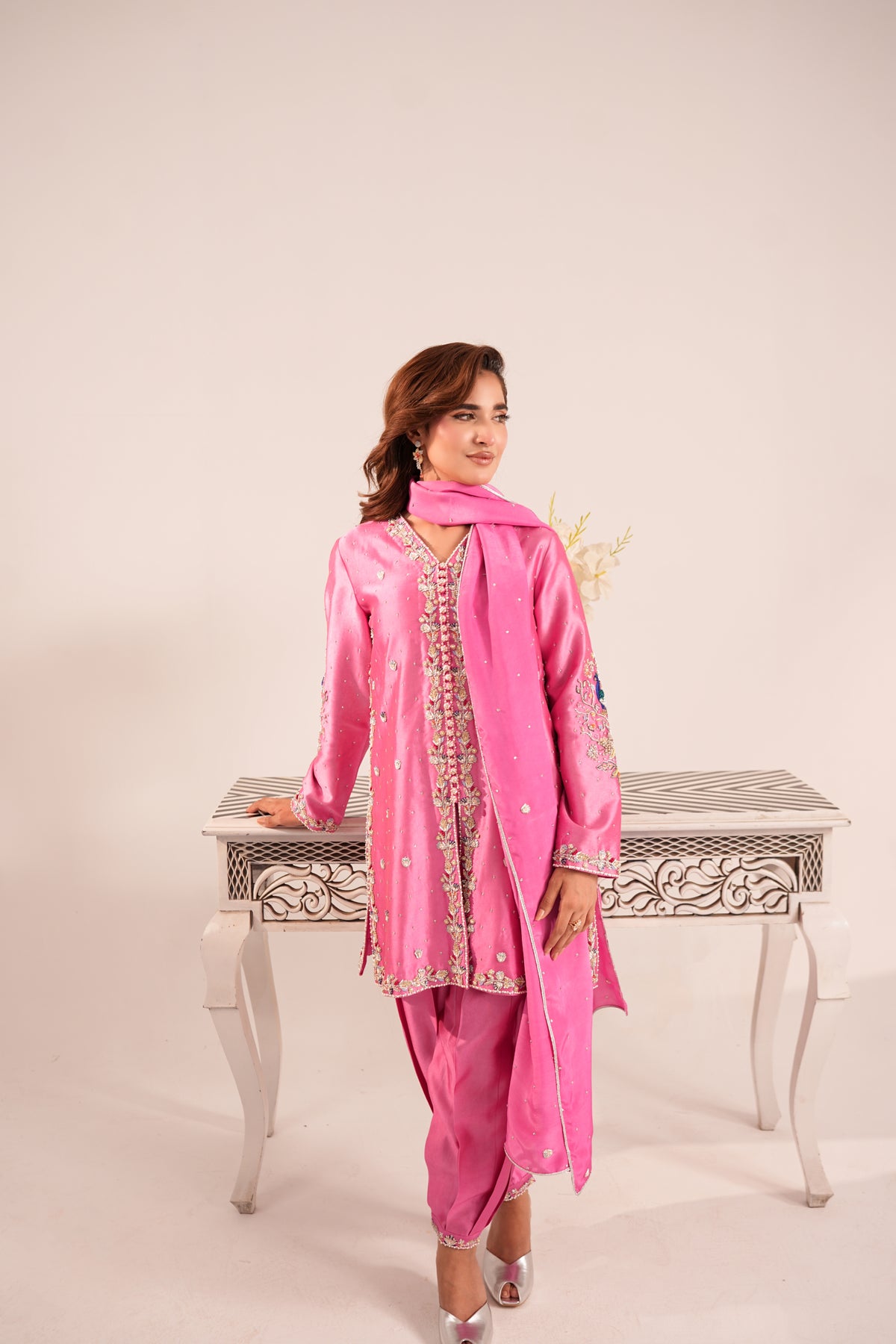 A woman modeling a bright pink traditional outfit with matching pants and a scarf, standing beside a decorative white table against a plain light beige studio background.