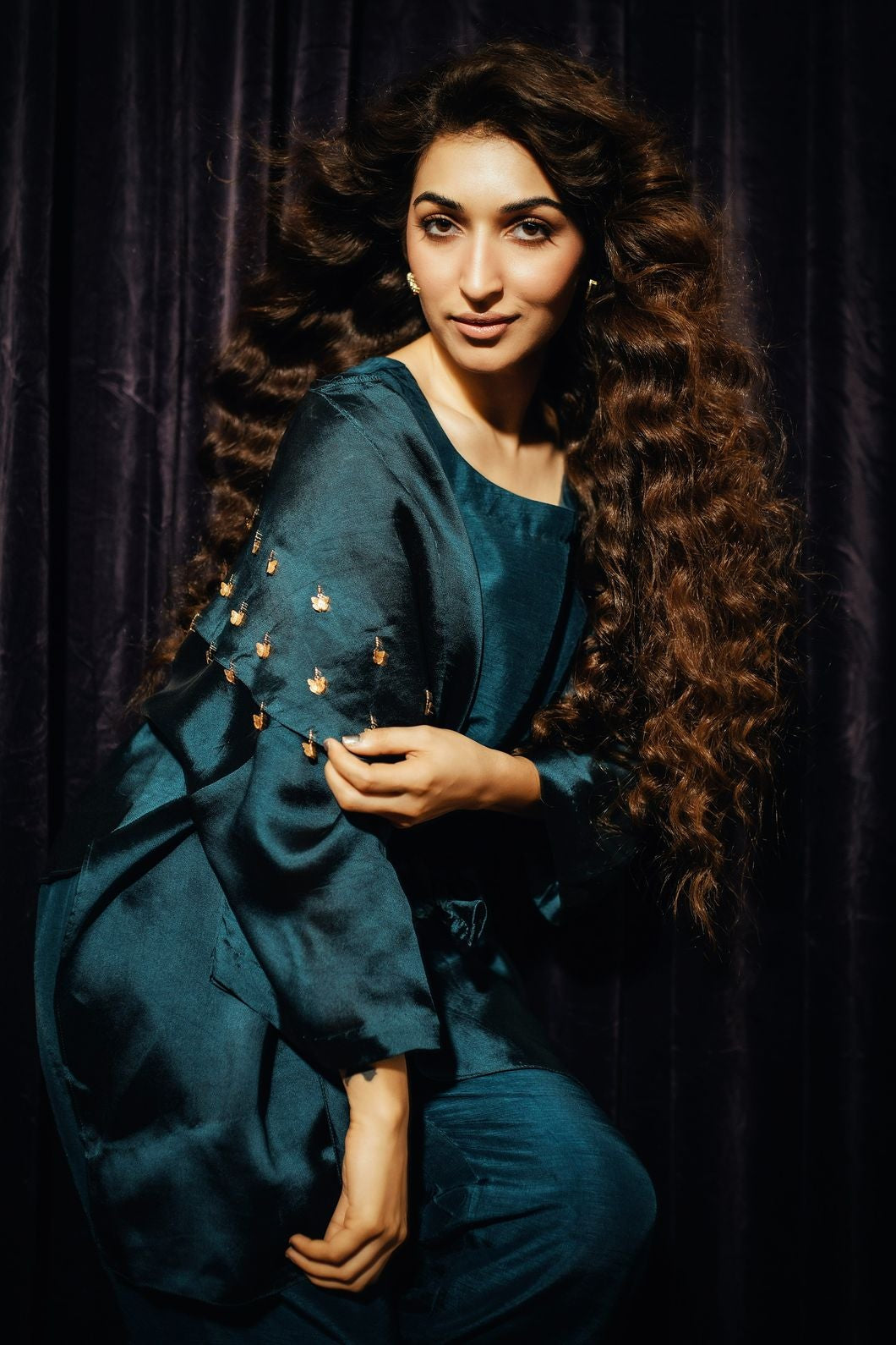 Woman in a teal outfit with gold embellishments against a dark background