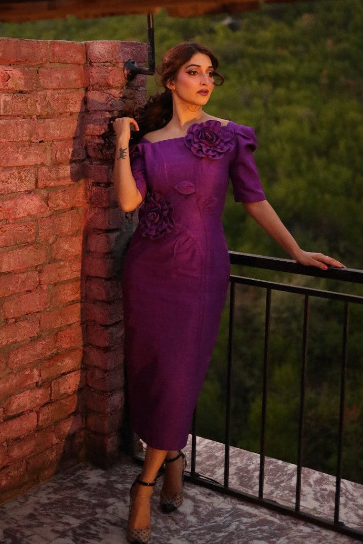 Woman in a purple dress standing on a balcony with a brick wall and greenery in the background