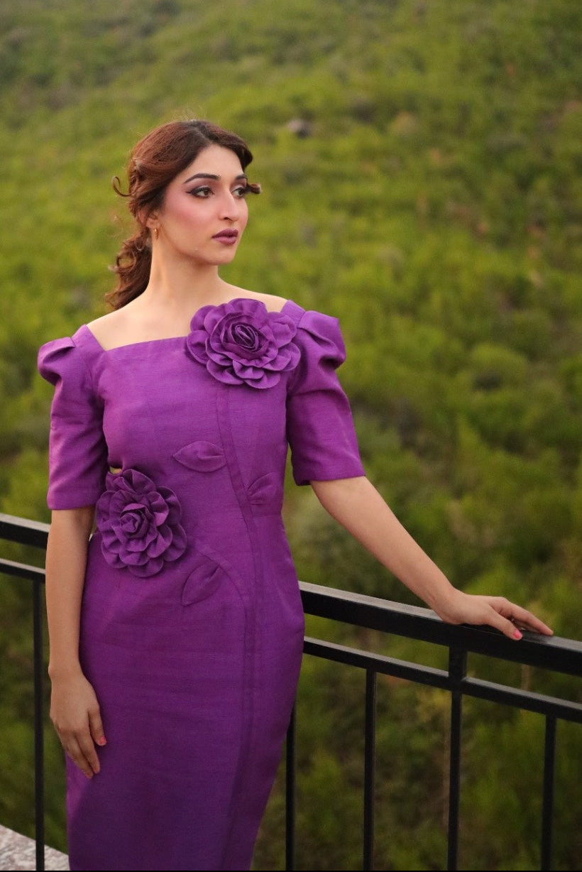 Woman in a purple dress with floral details standing against a green landscape.